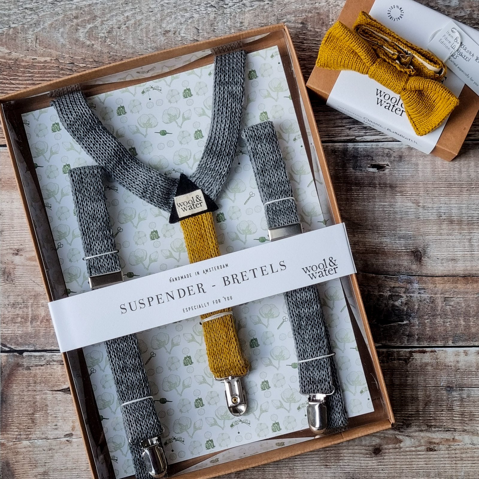 A pair of grey knitted suspenders with butterscotch yellow back in their Wool & Water packaging