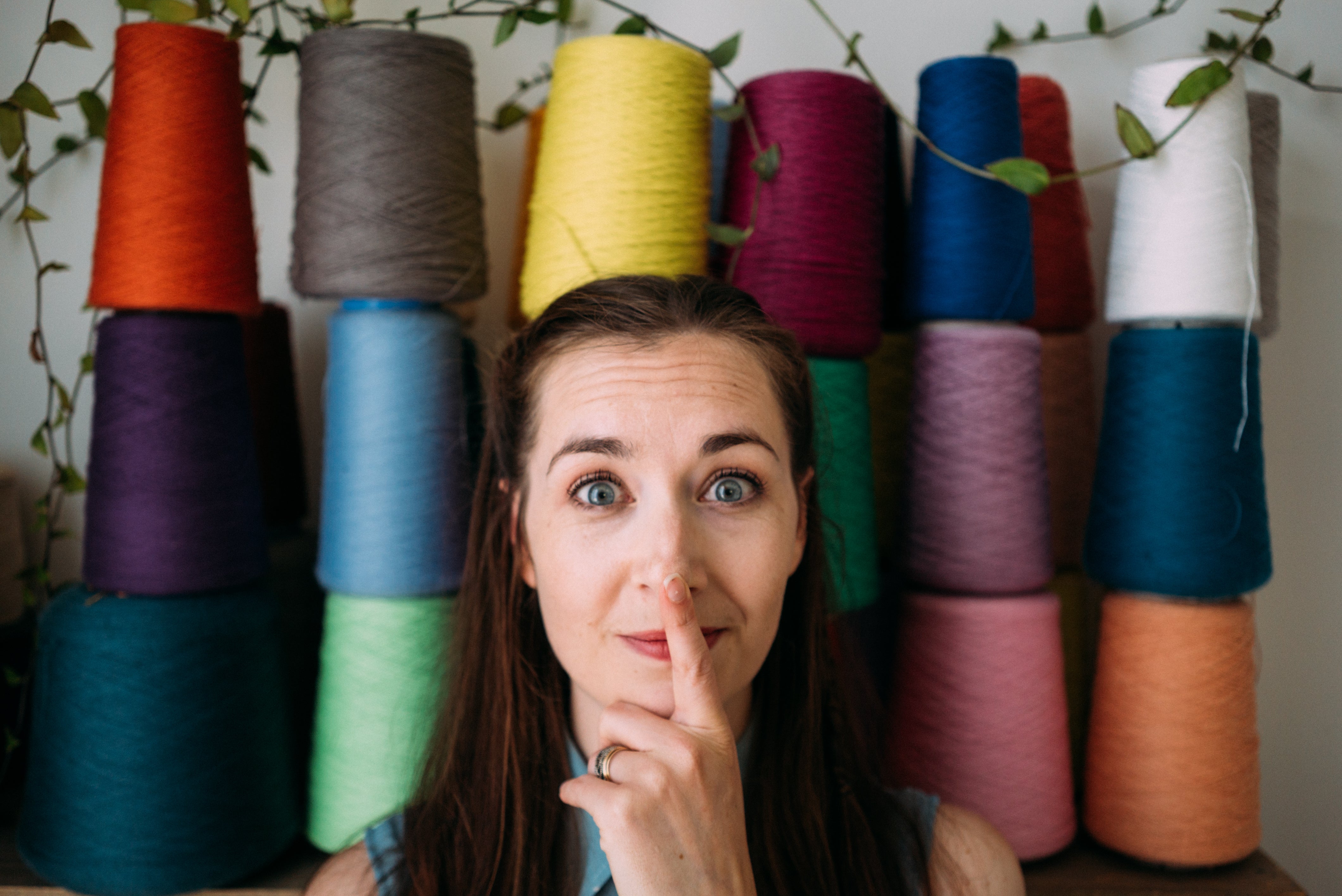 A white woman, Alice, in her 30's stands before a wall of multi-coloured yarn cones with her finger on her nose